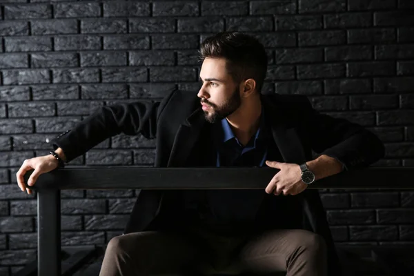 Fashionabla ung man sitter på bänken mot mörka tegelvägg — Stockfoto