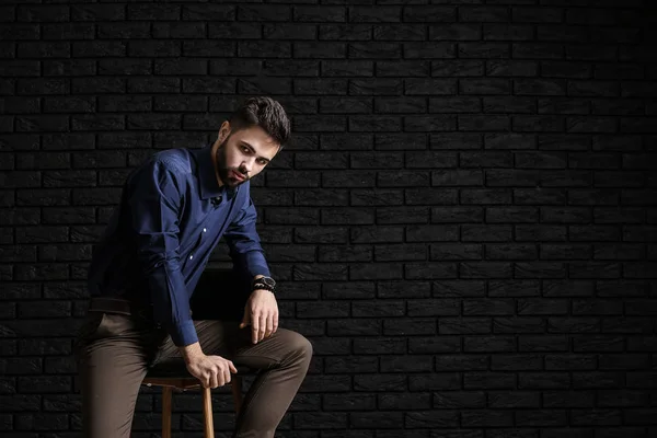 Jovem elegante sentado na cadeira contra a parede de tijolo escuro — Fotografia de Stock