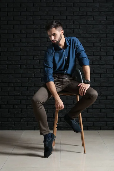 Модный молодой человек, сидящий на стуле у темной кирпичной стены — стоковое фото