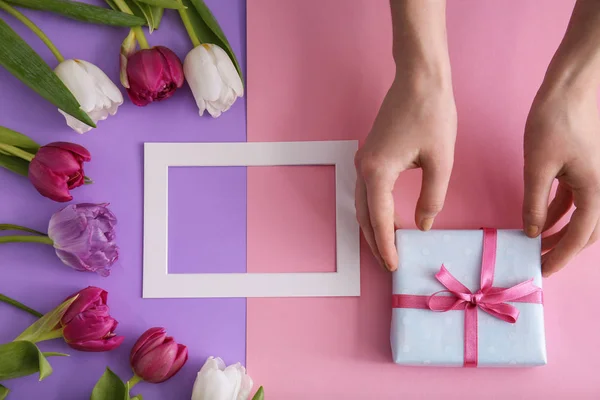 Female hands with gift box, beautiful flowers and frame on color background — Stock Photo, Image