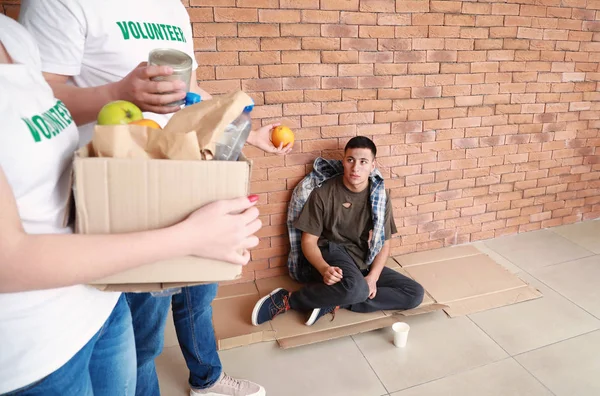 Voluntarios con comida y pobre hombre sentado cerca de la pared de ladrillo —  Fotos de Stock