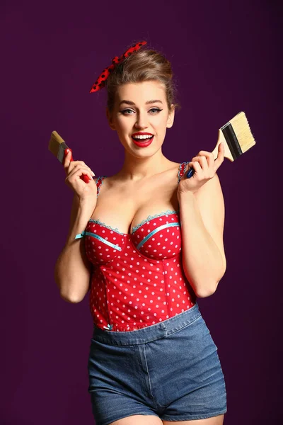 Beautiful pin-up woman with paint brushes on color background — Stock Photo, Image