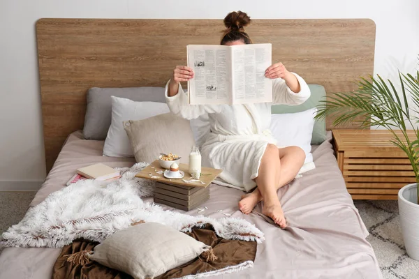 Jovem mulher lendo jornal e tomando café da manhã na cama — Fotografia de Stock