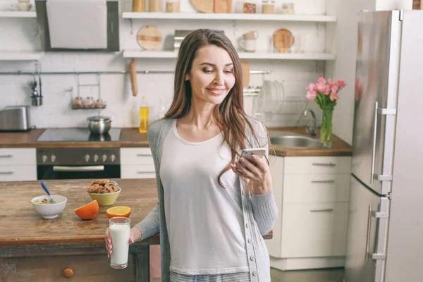 Vacker kvinna som dricker mjölk och använder mobil telefon i köket hemma — Stockfoto