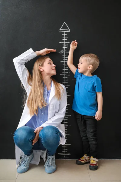 Médecin féminin avec petit garçon mesurant la hauteur près du mur sombre — Photo