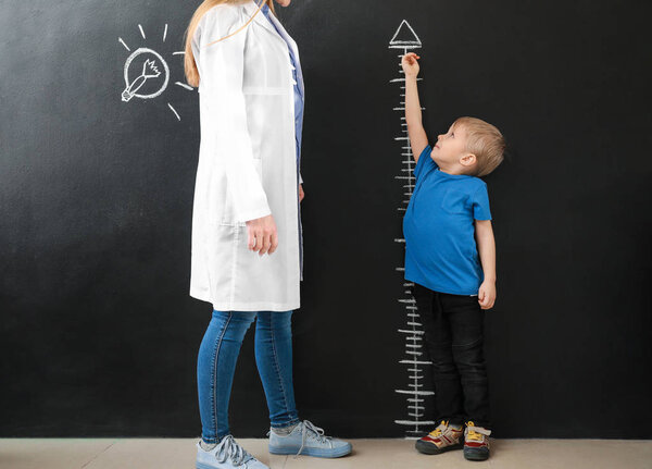 Female doctor with little boy measuring height near dark wall
