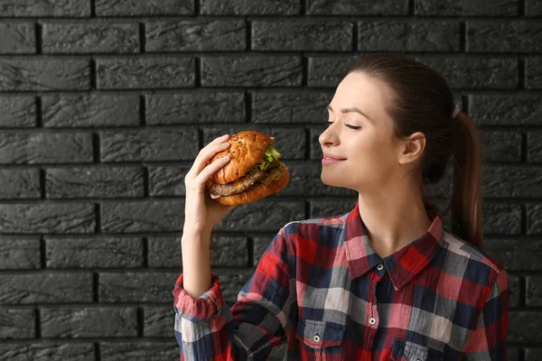 Mulher bonita com hambúrguer saboroso no fundo de tijolo escuro — Fotografia de Stock