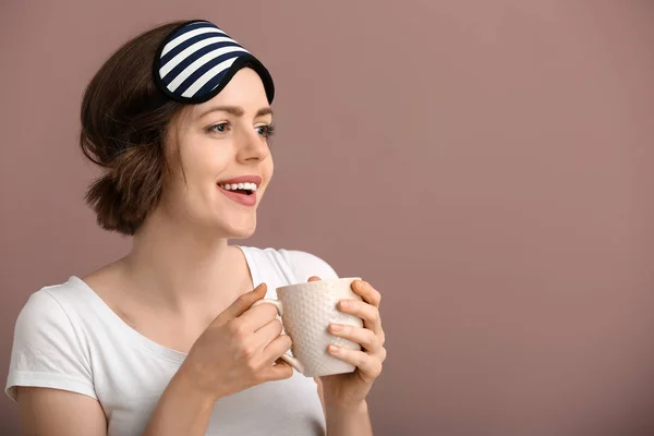 Happy young woman with sleep mask and cup of coffee on color background