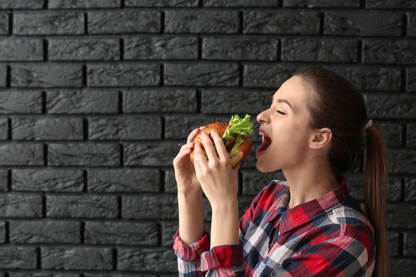 Bela jovem mulher comer saboroso hambúrguer no escuro tijolo fundo — Fotografia de Stock