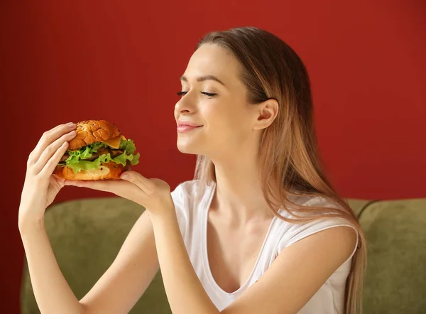 Beautiful young woman with tasty burger sitting on sofa — Stock Photo, Image