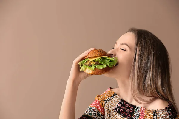 Schöne junge Frau isst leckeren Burger auf farbigem Hintergrund — Stockfoto