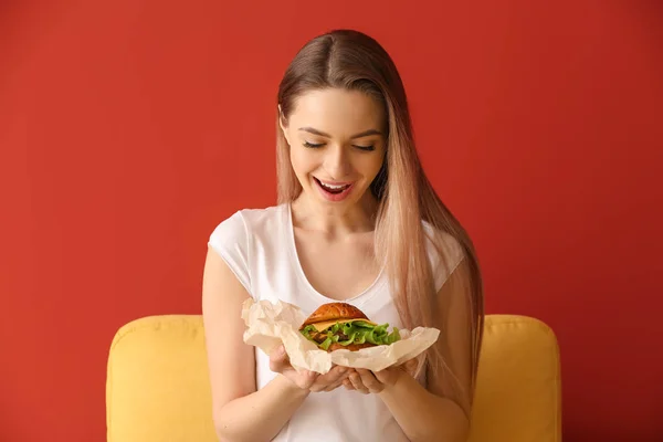Schöne glückliche junge Frau mit leckerem Burger auf farbigem Hintergrund — Stockfoto