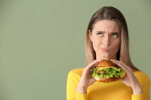 Jeune femme réfléchie avec hamburger savoureux sur fond de couleur — Photo