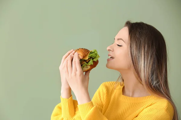 Красивая молодая женщина ест вкусный бургер на цветном фоне — стоковое фото