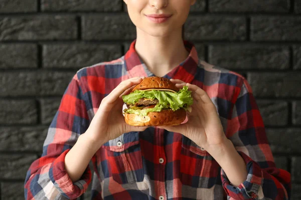 Schöne junge Frau mit leckerem Burger auf dunklem Backsteinhintergrund, Nahaufnahme — Stockfoto