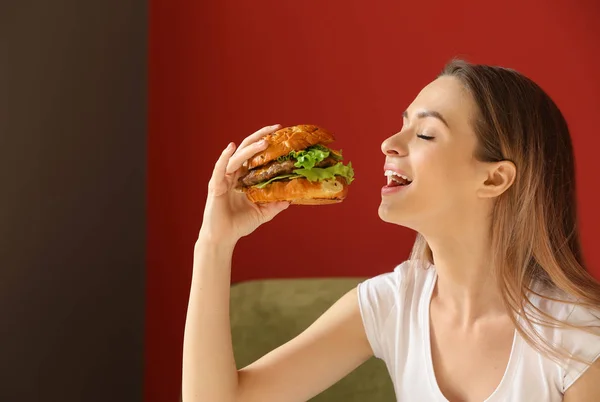 Beautiful young woman eating tasty burger indoors — Stock Photo, Image