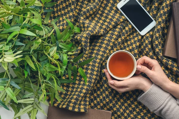 Female hands with cup of hot tea and mobile phone on plaid — Stock Photo, Image