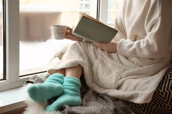 Mujer joven con taza de té caliente libro de lectura en alféizar de la ventana — Foto de Stock