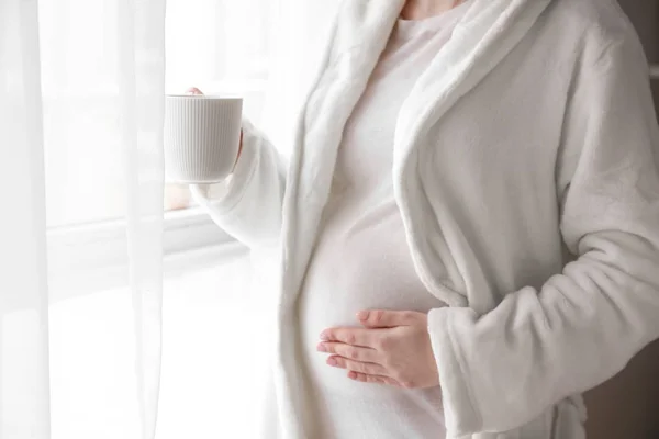 Těhotná žena s šálkem horkého čaje stojící poblíž okna doma — Stock fotografie