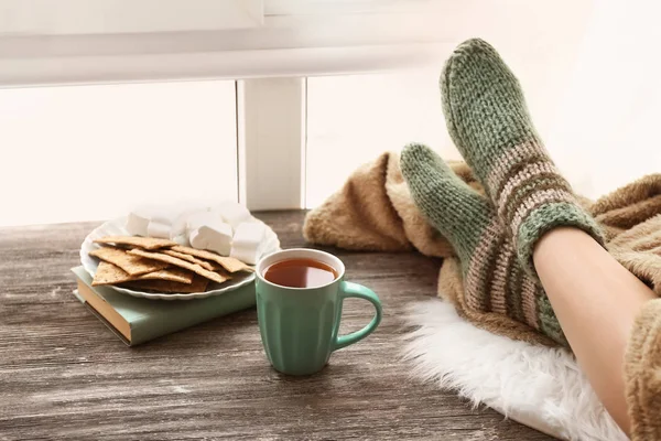 Kopp te med ben av ung kvinna på träbord nära fönster — Stockfoto