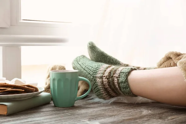 Cup of hot tea with legs of young woman on wooden table near window — Stock Photo, Image