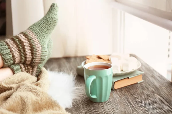 Taza de té caliente con patas de mujer joven en la mesa de madera cerca de la ventana — Foto de Stock