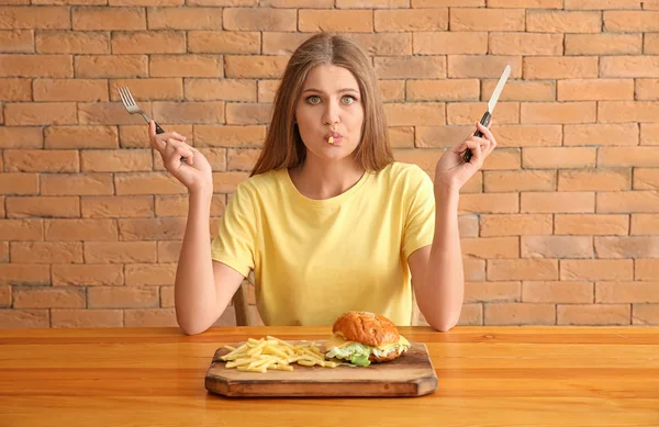 Mulher engraçada comer hambúrguer saboroso com batatas fritas à mesa — Fotografia de Stock
