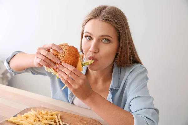 Mujer joven comiendo sabrosa hamburguesa con papas fritas en la mesa —  Fotos de Stock