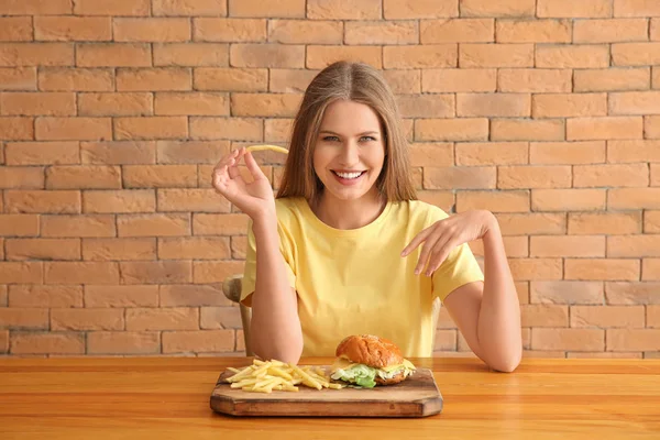 Junge Frau isst leckeren Burger mit Pommes am Tisch — Stockfoto