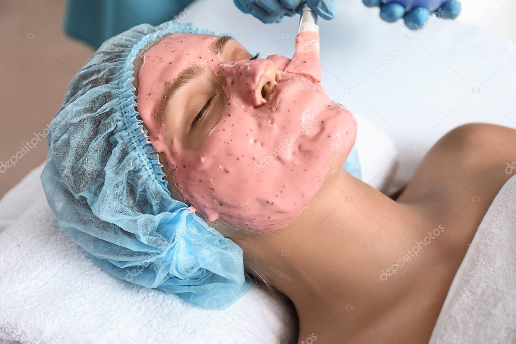 Cosmetologist applying alginate mask on young woman's face in beauty salon