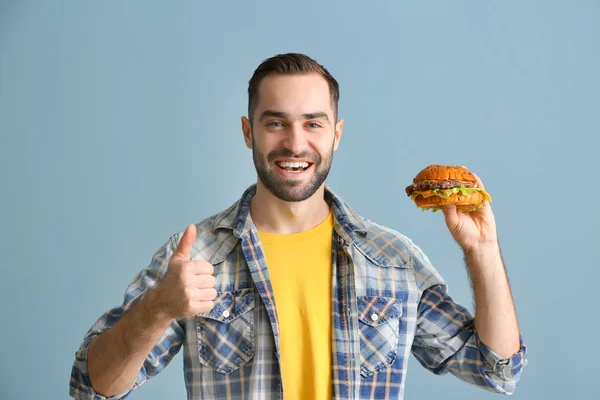 Mann mit leckerem Burger zeigt Daumen nach oben auf farbigem Hintergrund — Stockfoto
