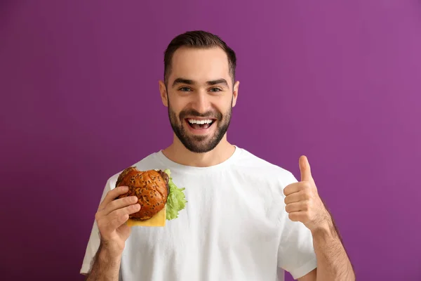 Hombre con sabrosa hamburguesa mostrando el pulgar hacia arriba en el fondo de color — Foto de Stock