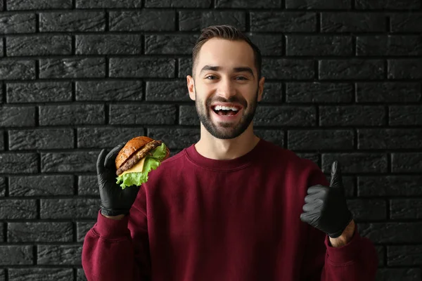 Uomo con gustoso hamburger mostrando pollice su sfondo scuro — Foto Stock