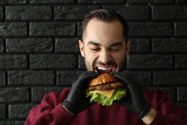 Mann isst leckeren Burger auf dunklem Hintergrund — Stockfoto
