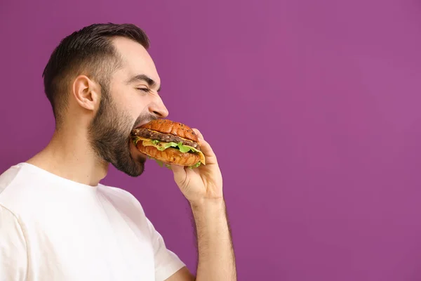 Mann isst leckeren Burger auf farbigem Hintergrund — Stockfoto