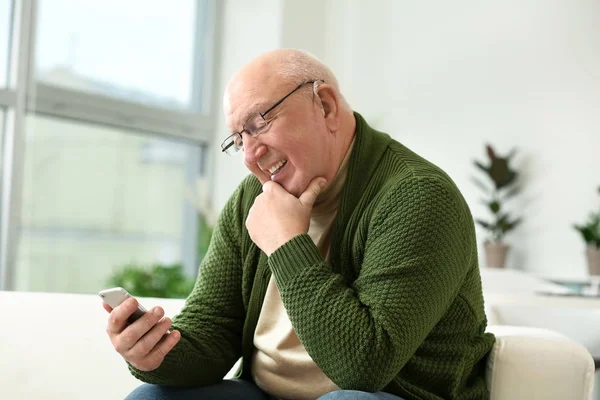 Starší muž s naslouchadlem doma pomocí mobilního telefonu — Stock fotografie