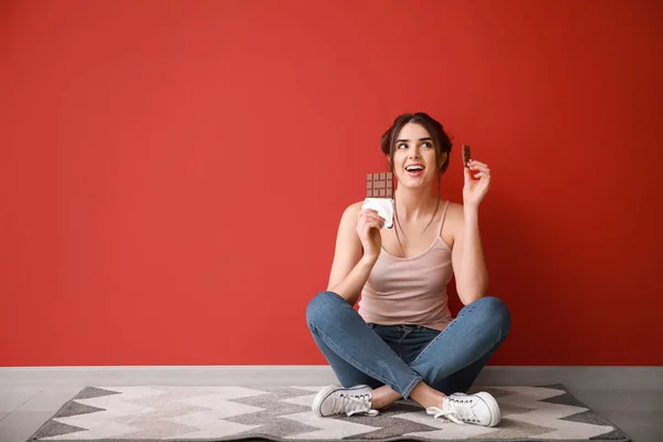 Beautiful young woman with tasty chocolate sitting on floor near color wall — Stock Photo, Image