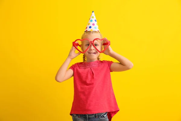 Funny little girl with big heart-shaped glasses and party hat on color background. April fools' day celebration — Stock Photo, Image