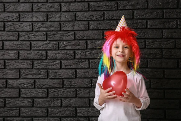 Funny little girl in bright wig and with balloon on dark background. April fools' day celebration — Stock Photo, Image