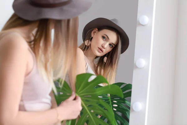 Beautiful woman with green tropical leaf looking in mirror indoors — Stock Photo, Image