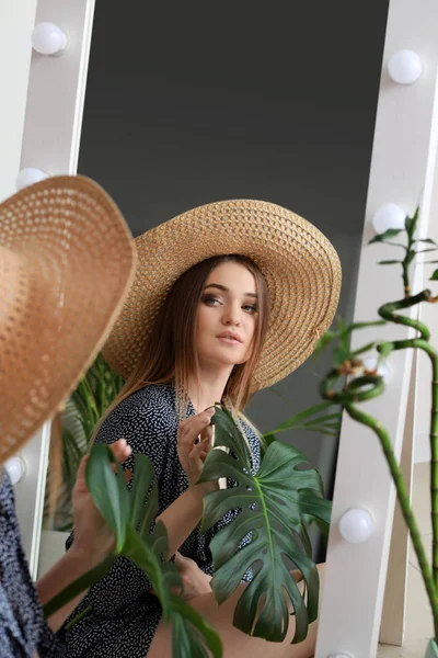 Beautiful woman with green tropical leaf looking in mirror indoors — Stock Photo, Image