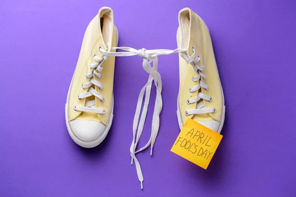 Shoes with tied together laces and sticky note with text APRIL FOOL'S DAY on color background