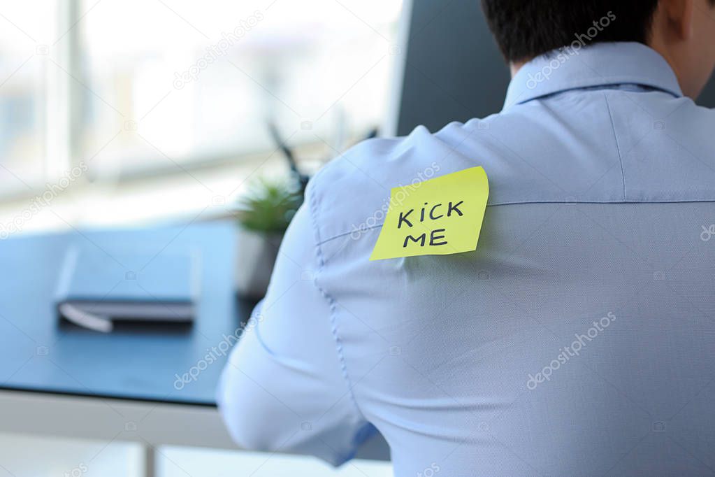 Sticky note with text KICK ME on back of man working in office. April Fools' Day prank