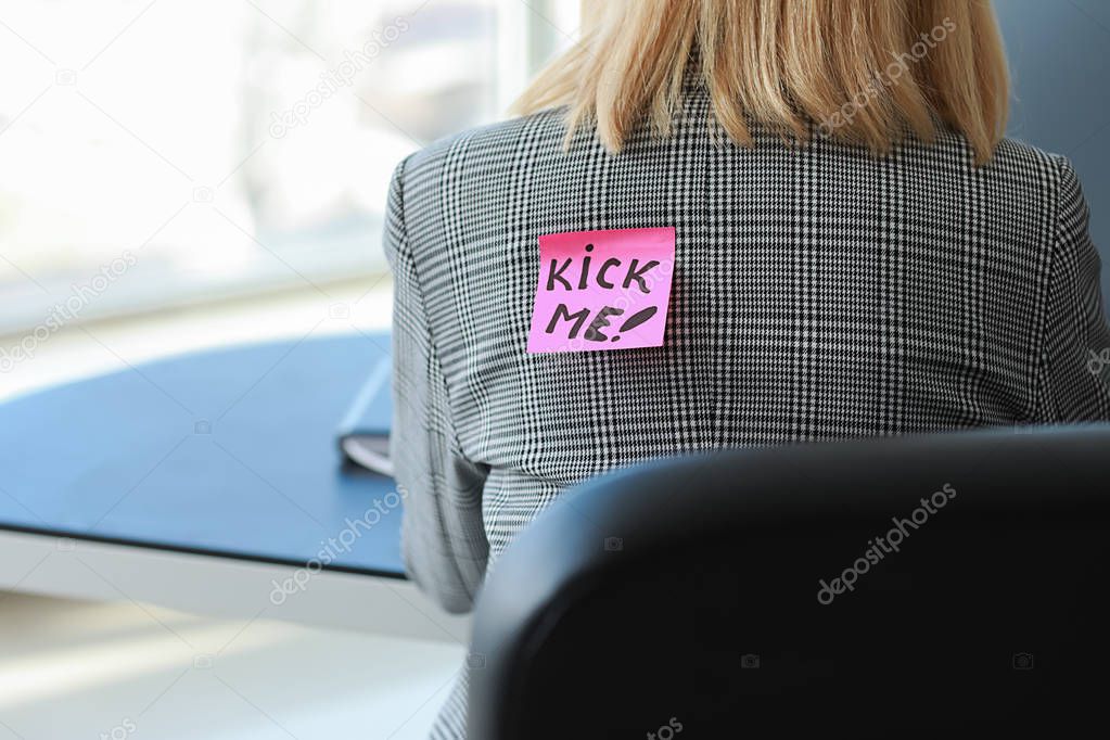 Sticky note with text KICK ME on back of woman working in office. April Fools' Day prank