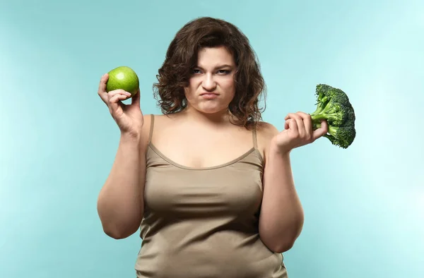 Displeased overweight woman with apple and broccoli on color background. Weight loss concept