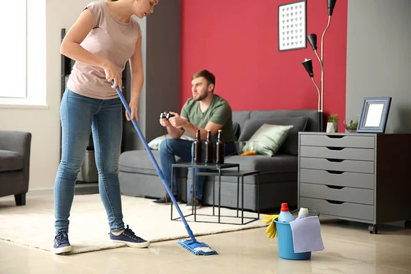 Wife cleaning floor while her lazy husband resting on couch at home
