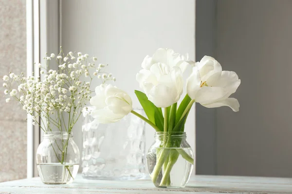 Jars with beautiful flowers on table near window — Stock Photo, Image