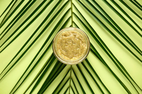 Jar of sugar scrub with tropical leaf on color background