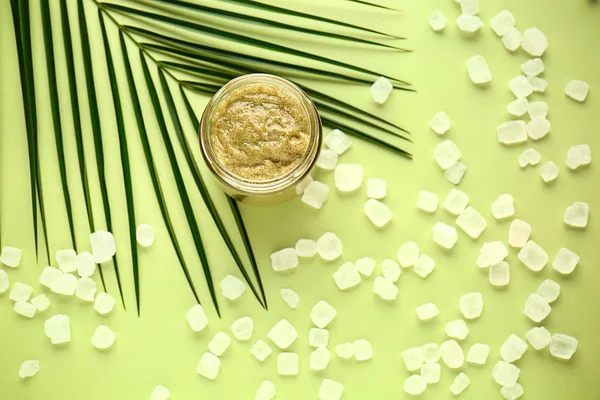 Jar of sugar scrub with tropical leaf on color background