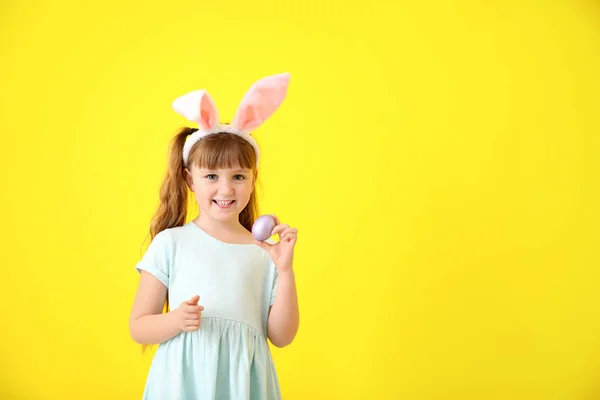 Симпатична маленька дівчинка з пасхальним яйцем і вухами кролика на кольоровому фоні — стокове фото